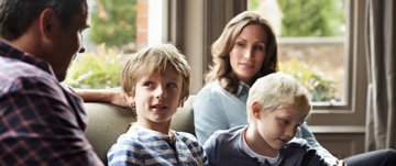 Mother, father, and two young children sitting together on the sofa, talking about chronic illness and how it will affect the family's future