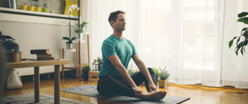 A man sits on a yoga mat in a cozy living room, surrounded by natural light and comfortable furnishings.