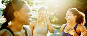 Three women joyfully laughing and smiling together under the warm sunlight, radiating happiness and friendship.