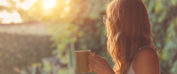 A woman enjoys a warm coffee mug as she gazes at a beautiful sunset, reflecting on self-isolation and personal growth.