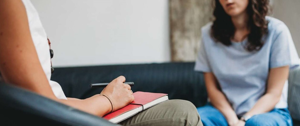  A woman sits on a couch with a notebook, accompanied by another woman, reflecting on her journey after a heart attack.